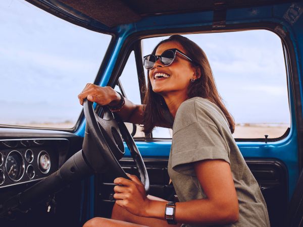 A smiling young woman wearing sunglasses sits behind the steering wheel of a vintage blue truck, laughing joyfully on a sunny day.