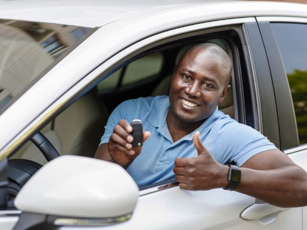 Black skinned guy seated in car with half open door showing thumbs up with car key in other hand